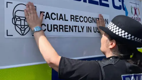 BBC A female police officer dressed in a dark uniform and round hat is sticking a sign with the words Facial Recognition Currently in Use on the side of a van.