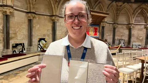 A woman standing inside Lincoln Cathedral holding two framed documents, one with handwritten text and the other with faint markings. There are arched columns in the background.
