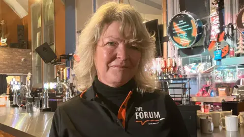 A head-and-shoulders portrait of Allison McKay standing in front of a bar. She has shoulder-length blond hair and green eyes. She is wearing a black turtleneck jumper under a black polo shirt with the logo of The Forum with white and orange detailing.