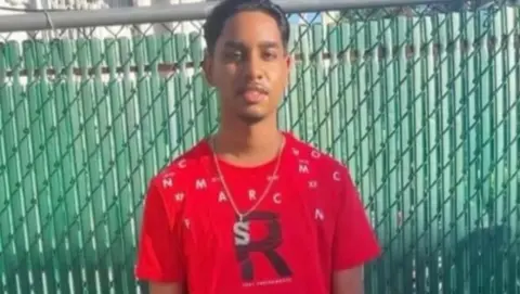 Family Shawn Seesahai wearing a red T-shirt and neck chain. He is standing in front of a green fence.