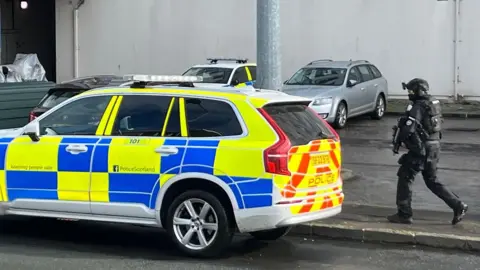 An armed police officer walking near a marked police car. There are other cars parked nearby, including another police car.