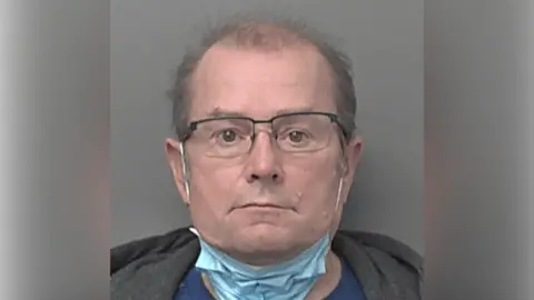 A police custody photo of Darren Richardson. He has short, sparse brown hair and is wearing black-rimmed glasses and a face mask under his chin, with the straps around his ears. He is also wearing a blue T-shirt and a grey hoodie. The wall behind him is grey.