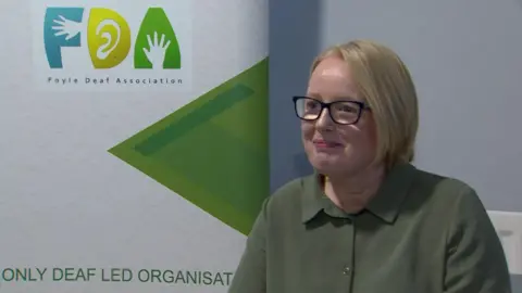 A woman with short blonde hair and glasses sits smiling looking to the left of the camera. She is wearing a green top and in the background is a standing poster board for Foyle Deaf Association