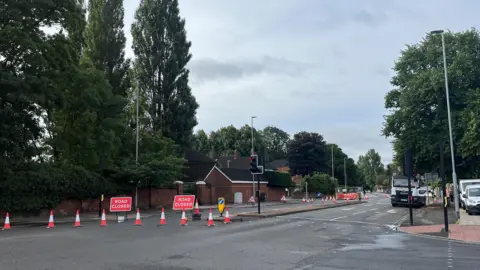 BBC/ Louise Hobson A grey tarmac road with two lanes on either side is closed on one side. Orange and white cones are placed diagonally across the road and there are two red signs in place that read: "Road closed." There is a pedestrian crossing and white vans parked on the road, which is bordered by tall trees.