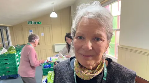 John Devine/BBC Carol Banks has short grey hair swept across her forehead, she is wearing a woollen fawn coloured top, with a yellow and pink scarf around her neck. behind her two women pack shopping bags with supplies in the food bank.