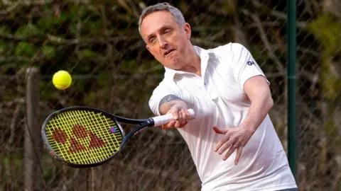 PA Media Alex Cole-Hamilton, who has short grey hair, swings a tennis racket at a ball on an outdoor court  