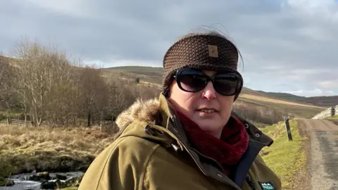Sam Wood is in her 40s and wearing a warm jacket, red scarf and hat and sunglasses. Behind her is a single track road with a river to its left and hills behind it 