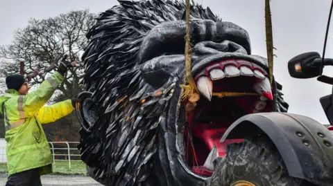 The British Ironwork Centre A large roaring gorilla head made of iron, being unloaded off of a machine, attached to ropes 