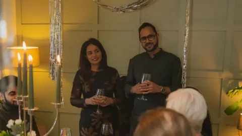 Your Pal Moments A woman and a man standing at the top of a table. They are both holding wine glasses and smiling at a woman sitting at the table.