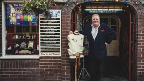 The World's Smallest Magic and Joke Shop Older photograph of Malcolm Norton stood outside his shop with the straitjacket on a mannequin. Mr Norton is clean shaven and smiling at the camera with his right arm resting on the mannequin. He is wearing a dark blue jacket and dark coloured trousers.