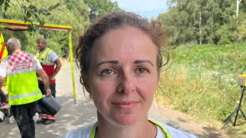 John Devine/BBC Vicky Best is looking straight at the camera with a neutral expression. She is standing in front of a grassy area and a number of officials in high-visibility vests are talking behind her.