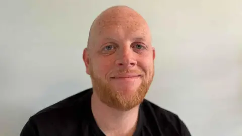 Jodie Halford/BBC Liam Smith is looking towards the camera with a light smile. He has a short ginger beard, and is wearing a black T-shirt.