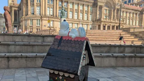 A sculpture of Snoopy the cartoon dog lying on a small wooden house, which is placed in front of Birmingham's Town Hall.