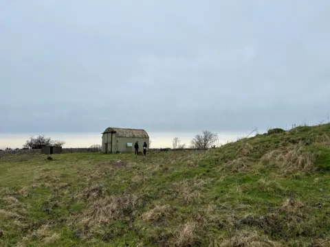 Two men stand in front of a metal hut in a field.
