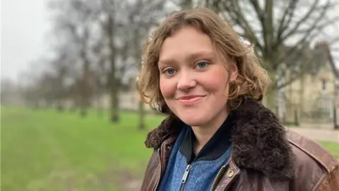 Stephen Huntley/BBC Fran Lusty smiling and standing in a park. She wears a brown leather jacket with a fake fur collar with a blue denim jacket underneath. She has shoulder-length brown hair which is slightly curly.