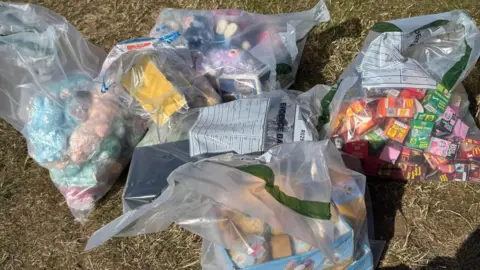 Plastic bags full of counterfeit toys are strewn on some dry grass. One of the bags has colourful boxes and another has colourful soft toys in it.