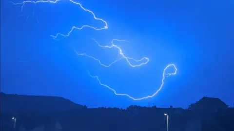 Serveral long lighting bolts across a pale blue night sky with dark hills and houses on the horizon