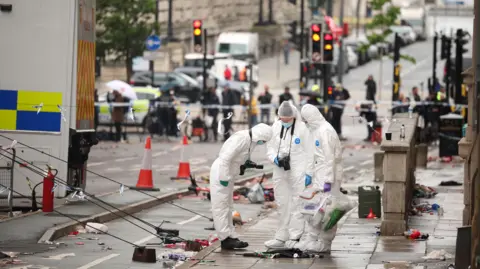 DAM VAUGHAN/EPA-EFE/Shutterstock Three people are wearing forensic suits with cameras around their necks. They are standing on a pavement with rubbish scattered around them. In the background cars are parked on the road.
