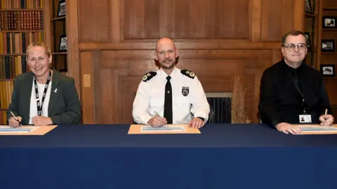 TVP Aileen O'Connor, dressed in a grey jacket and white shirt, Jason Hogg, dressed in his chief constable's uniform, and Ken Cooney, wearing a black shirt and tie and dark-rimmed glasses, signing the pledge. They all have pens in their hands.