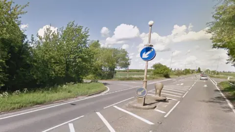 The junction of a main raid with two side roads, surrounded by trees and green trees. The grey asphalt is split by a crossing point with blue signs telling motorists to keep left. White road markings denote the turn-offs.