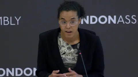 Kaya Comer-Schwartz, London's deputy mayor for policing and crime, speaking into a microphone at a London Assembly meeting. She has dark curly hair and glasses and is wearing a black blazer.