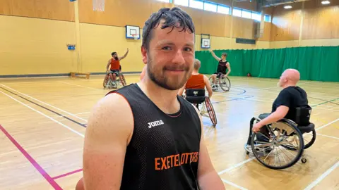 BBC Jack Davey smiling in the front view with four men playing wheelchair basketball behind him