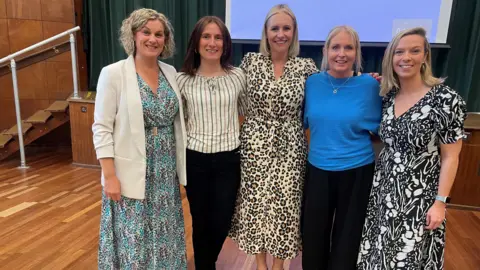 A group of five women stand in an assembly hall with their arms around each other. Three are wearing patterned dresses and two are wearing dark trousers and a top. All women are smiling into the camera.