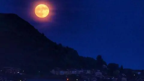 A full moon tinged pink/orange in a deep blue sky, with the silhouette of a mountainside and houses below in Tonypandy, Rhondda Cynon Taf