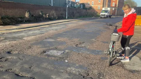 A picture of a woman wearing a red coat with black cycling leggings stood by the side of the road with her electric bike. The road is covered in potholes.