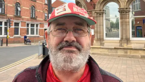 Steve Jones/BBC Carl Jones, who has a grey beard and glasses. He is wearing a red Liverpool FC cap.
