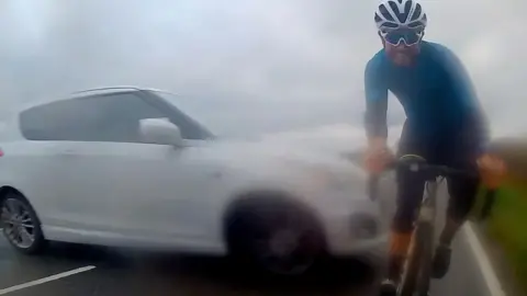 A white car slides sideways across a road, narrowly missing a cyclist who is facing the camera.