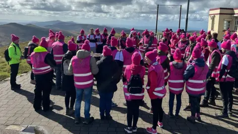 A large crowd of people wearing pink coats and pink woolly hats standing next to the cafe at the top of Snaefell, looking out at the hills on a sunny day.