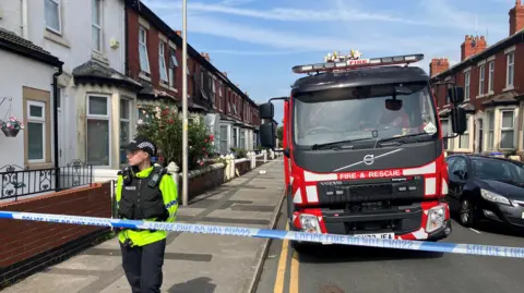 BBC/Yunus Mulla A police officer stands outside the scene of a house fire as a fire truck is seen behind him on the street. Police tape crosses the front of the pic. The houses are Victorian in style with big bay windows. 