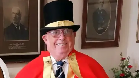 Terrence Mullett Man wearing a black top hat with a yellow buckled ribbon across the middle and is wearing a red cloak over a white shirt and blue stripy tie with a golden clasp fashioning the cloak.