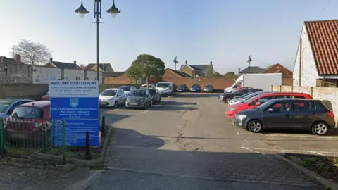 A Google street view image of the car park. There is a blue-and-white car park sign at the entrance to the ground-level car park, which has spaces marked in white paint. There are cars in some of the spaces.