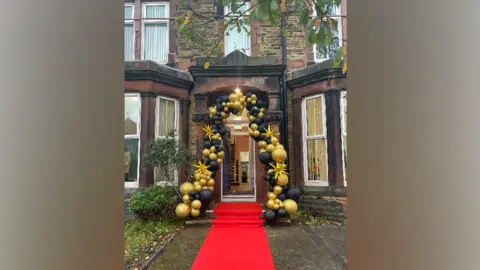 Sefton Council Black and gold balloons frame a Victorian doorway of a house, a red carpet extends from the front door to the front garden, the front door is open