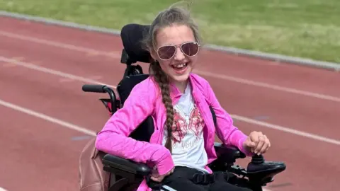 PA Media Tammy is sat in a black wheelchair on an athletics track which is red. She has long brown hair in a plait, and is wearing a white top with a pink butterfly on and a pink jacket. She is looking at towards the viewer smiling, and is wearing sunglasses.