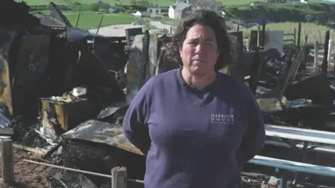 BBC Ms Costin stood in front of the wreckage wearing a navy blue jumper. She has dark curly shoulder-length hair.