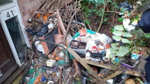 Rubbish at the door of a property. There is garden waste, plant pots, bottles and black bags. 