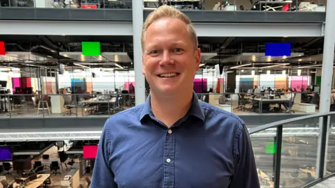 A man smiles and looks at the camera as he stands on a balcony in the foyer of the BBC building in Cardiff. He has short blond hair and is wearing a dark blue shirt.