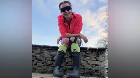 Laura Stenton-Slater A woman sits on a bench, looking down the camera. She has brown hair tied back in plaits, and wears a fluorescent pink running jacket with a hood, large sunglasses, black running shorts and black wellington boots. Her knees are strapped up with black support strips. 