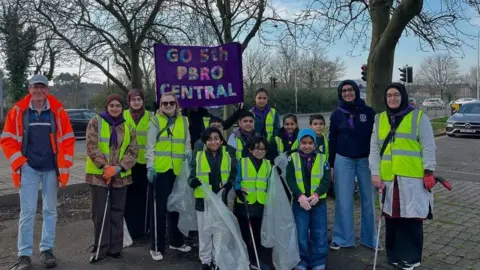 Shama Akhtar Adults and children in hi-vis jackets hold bin bags and litter picking tools. They are all smiling at the camera. There's a banner behind them that says "Go 5th Pbro Central".