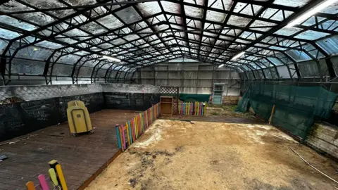 Cleethorpes Wildlife Rescue The interior of a large glass house. It has a glass-panelled roof and walls, upheld by a black complex steel structure. The glass panels look dirty. Most of the floor is covered with sand, but a raised area of wooden decking stands to the left, surrounded by a rainbow-coloured wooden fence.
