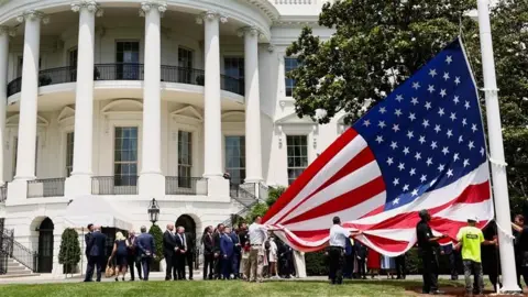 A US flag is held up by workers who are readying it up to be hung on a tall pole outside the White House as Trump and others look on.