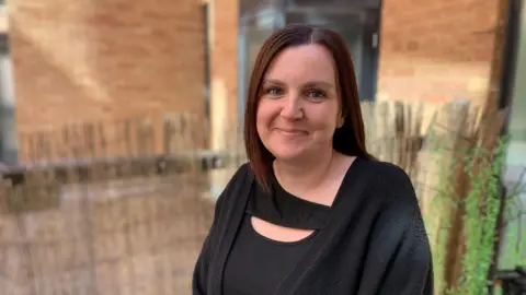A woman smiles at the camera. She is wearing a black top and black cardigan. She is standing outside.