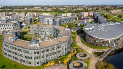 An aerial image of the University of York campus in Heslington. Many of the buildings are at least three storeys high and very modern in appearance. There is a lake next to one of the buildings with a walkway leading over it. Green trees and houses can be seen in the distance.