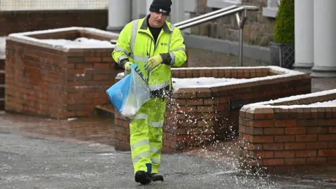 Pacemaker A man in yellow high-viz trousers and jacket and a black woollen hat throws salt out of a bucket on to a footpath