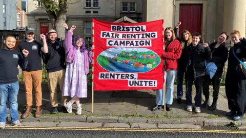 Bristol Fair Renting Campaigners posing for a photo around a red banner reading "Bristol Fair Renting Campaign - renters unite!". Most have a fist raised.