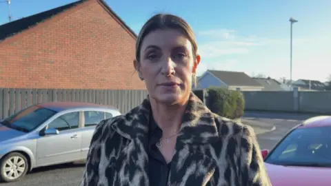 Linda Dillon. Tiene el cabello castaño oscuro recogido, lleva un abrigo con estampado de leopardo, una camisa negra debajo y un collar plateado con un corazón y una "L". Está de pie junto a un coche rojo con un coche plateado aparcado y casas al fondo.