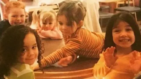 Five young girls look at the camera while sitting around a table at McDonald's in 1997. The picture has an orange tint. The girl on the right is waving at the camera and beaming, while the girl next to her is Alice Connor and is leaning across the table. They are made up to look like young versions of the Spice Girls. 